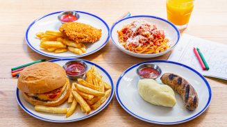 A table with four plated meals including a cheeseburger with fries, fried chicken with fries, spaghetti with tomato sauce and cheese, and grilled sausage with mashed potatoes.