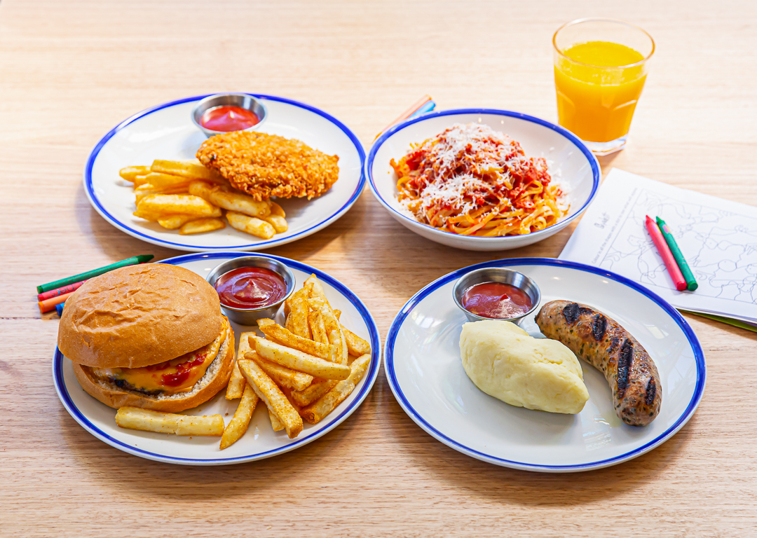 A table with four plated meals including a cheeseburger with fries, fried chicken with fries, spaghetti with tomato sauce and cheese, and grilled sausage with mashed potatoes.